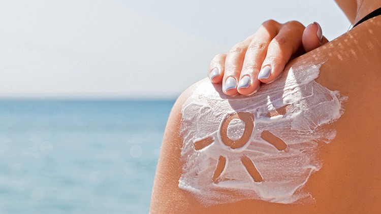Eine Schulter am Strand, darauf Sonnencreme mit einem Sonnensymbol, eine Hand mit hell lackierten Nägeln berührt die Creme.