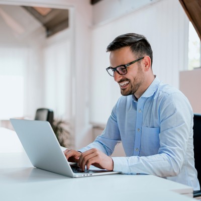 Person in blauem Hemd arbeitet an einem Laptop in einem hellen Büro mit Fenstern und Holzdetails.