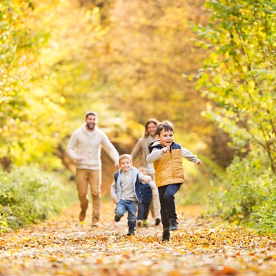 Eine Familie spaziert im Herbst durch einen Waldweg mit buntem Laub, zwei Kinder rennen spielend vorne weg.