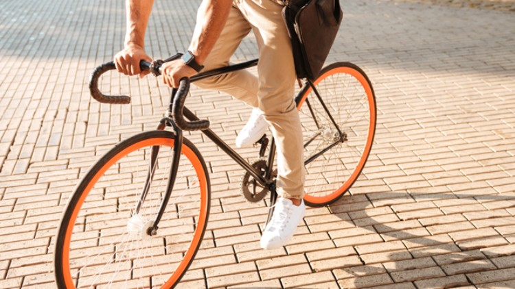 Person in heller Hose und weißen Schuhen fährt auf einem schwarzen Fahrrad mit orangefarbenen Reifen über gepflasterten Boden.