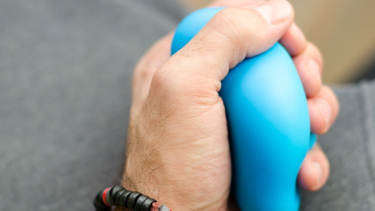 Eine Hand hält einen blauen, ovalen Stressball, im Hintergrund ist ein Teil von grauer Kleidung sichtbar.