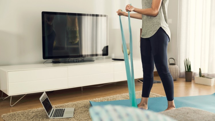 Person steht auf einer blauen Yogamatte, hält ein Fitnessband gespannt, im Hintergrund Laptop, Fernseher und heller Wohnbereich.