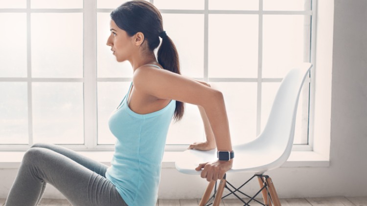 Person macht Fitnessübung auf einem weißen Stuhl vor einem großen Fenster, trägt Sportkleidung und eine Smartwatch.