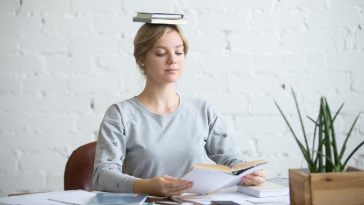 Person in grauem Pullover hält ein geöffnetes Buch, während mehrere Bücher und Papierstapel auf dem Tisch liegen.