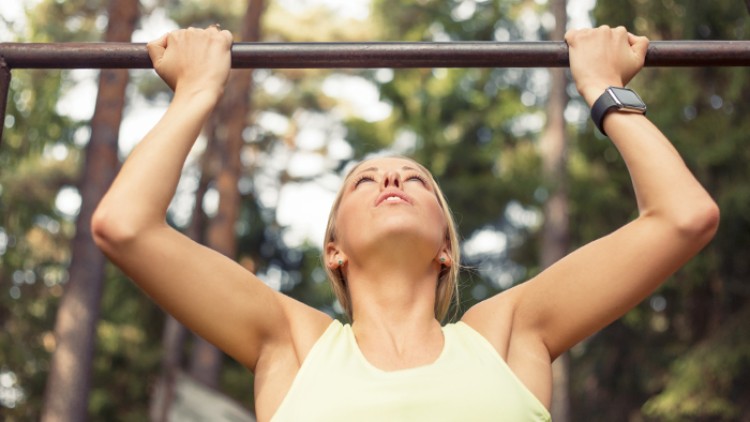Person in gelbem Sporttop macht Klimmzüge an einer Stange im Freien, umgeben von Bäumen, mit Fitnessuhr am Handgelenk.