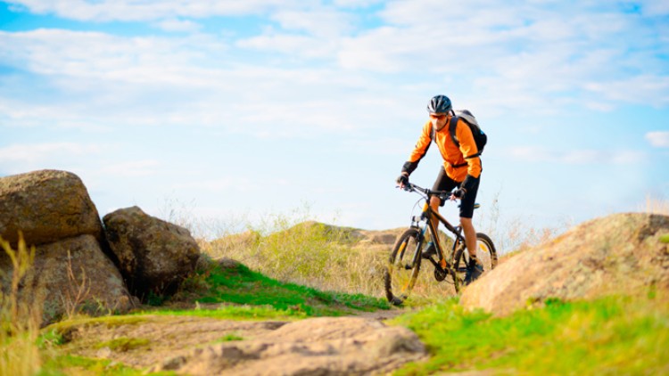 Person mit orange-schwarzer Kleidung und Fahrradhelm fährt auf einem Mountainbike durch eine felsige Landschaft mit blauem Himmel.