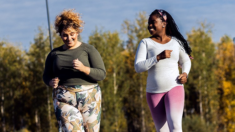 Zwei Personen joggen in sportlicher Kleidung auf einem Weg vor herbstlich gefärbten Bäumen und einem blauen Himmel.