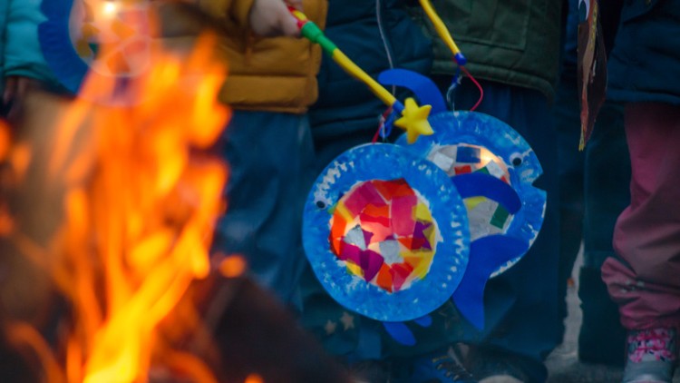 Mehrere bunte Laternen mit transparenten Papierabschnitten und Stern-Dekorationen werden von Kindern nahe einem Feuer getragen.