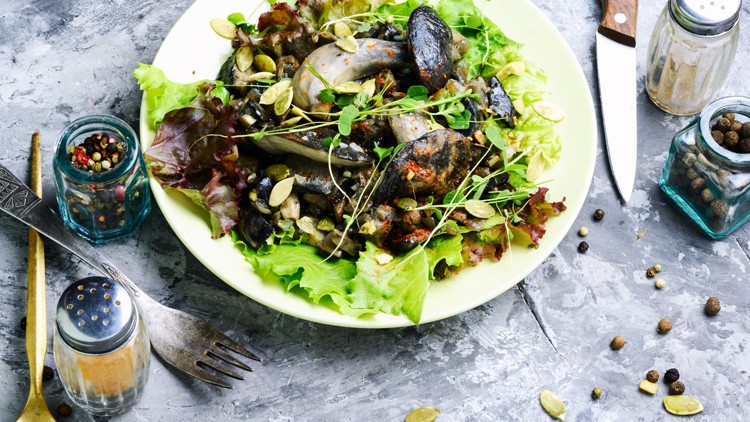 Ein Teller mit gemischtem Salat, gerösteten Kürbiskernen, gegrillten Pilzen und Sprossen, umgeben von Gewürzstreuern und Besteck.