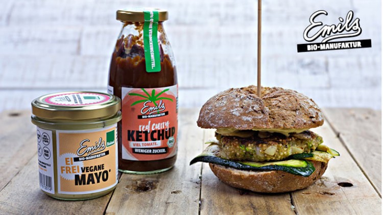 Ein rustikaler Burger mit Zucchinischeiben steht neben einem Glas „Ei frei vegane Mayo“ und einer Flasche „Red Curry Ketchup“.