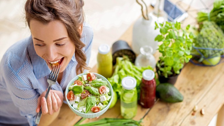 Person hält eine Schale mit Salat aus Tomaten, Feta und Basilikum, auf einem Tisch liegen Gemüse, Kräuter und Getränke.