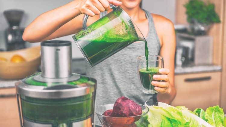 Person gießt grünen Gemüsesaft aus einem Mixerkrug in ein Glas, auf dem Tisch liegen Salat, Süßkartoffeln und Rote Bete.