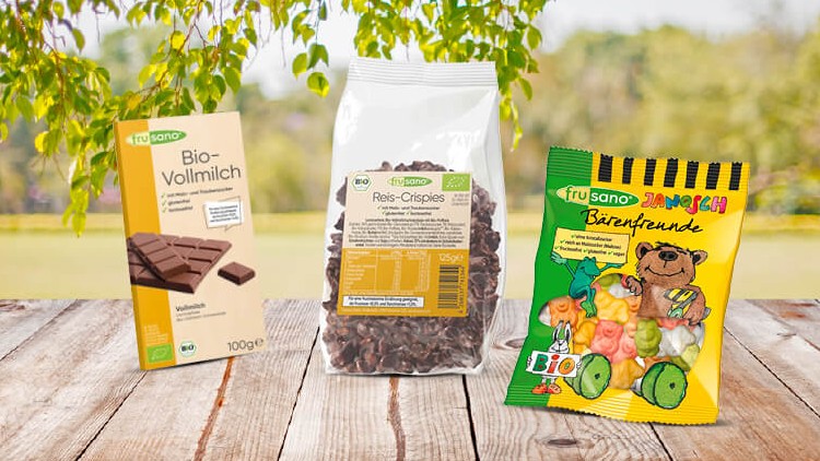 Drei biologische Produkte von frusano, Schokolade, Reis-Crispies und Janosch Bärenfreunde, stehen auf einem Holztisch im Freien.