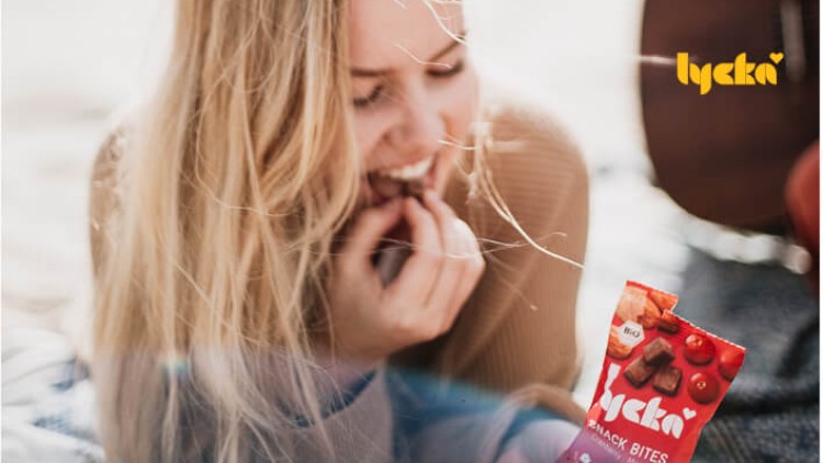 Person hält eine rote Packung „lycka Snack Bites Bio“ in der Hand, im Hintergrund verschwommene Details und „lycka“ Schriftzug.