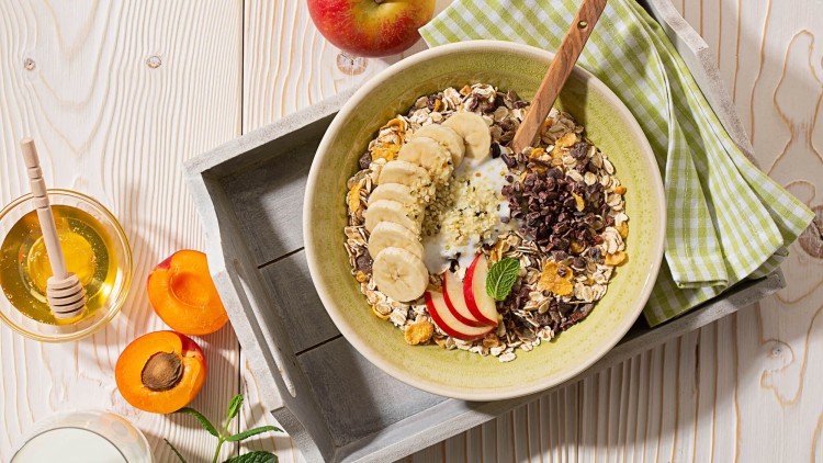 Eine grüne Schale mit Müsli, Bananenscheiben, Apfelstücken, Joghurt und Minze, daneben Honig, Aprikosen und ein Glas Milch.