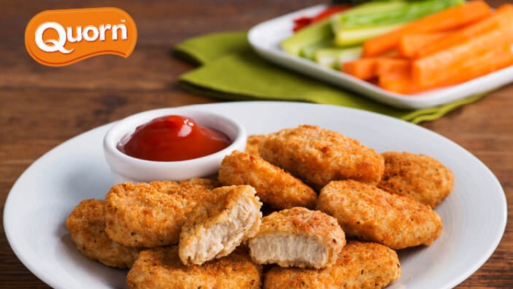 Auf einem Teller liegen goldbraune Nuggets mit einer Schale Ketchup, im Hintergrund Gemüsesticks, oben links der Schriftzug Quorn.
