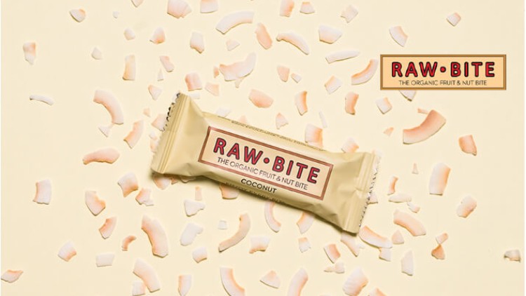 Ein beige verpackter Kokosnuss-Riegel mit der Aufschrift RAW BITE liegt auf beigem Hintergrund mit verstreuten Kokoschips.