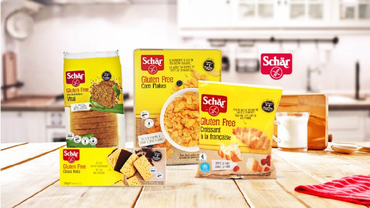 Drei glutenfreie Schär-Produkte auf einem Holztisch in einer Küche, darunter Vitalbrot, Cornflakes und Croissants à la française.
