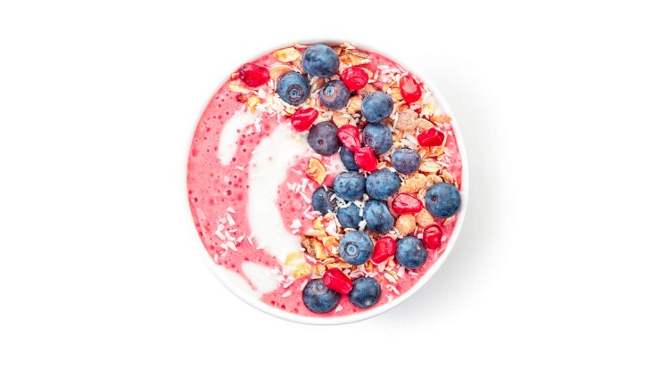 Eine Schale mit rosa Smoothiebasis, darauf Blaubeeren, Granatapfelkerne, Nüsse, Kokosflocken und Müsli, auf weißem Hintergrund.