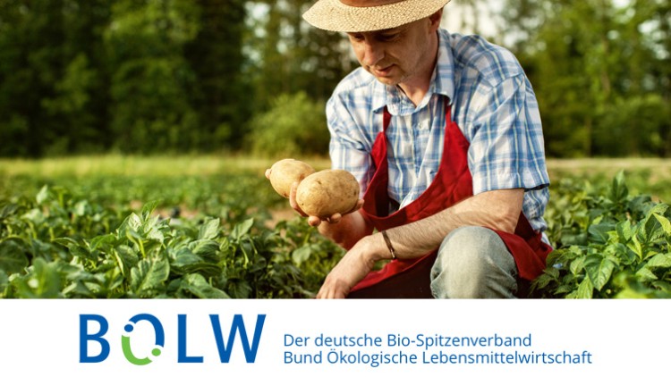 Person mit Strohhut und roter Schürze hält zwei Kartoffeln in einer grünen Feldlandschaft, darunter BÖLW-Logo mit Text.