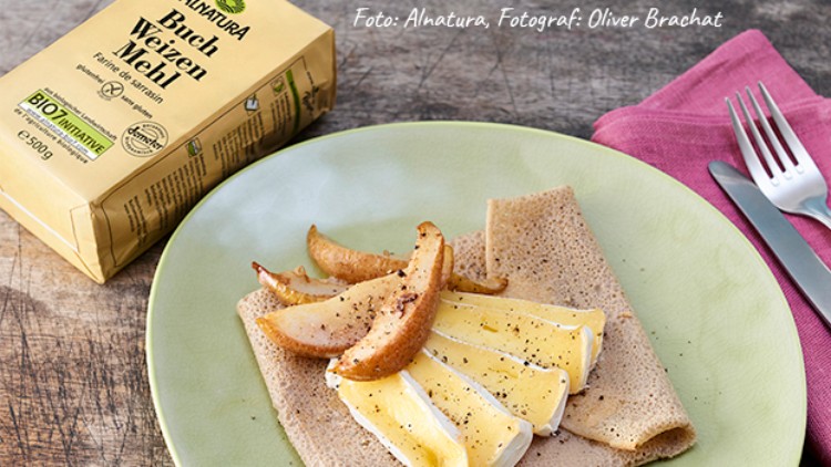 Ein Teller mit Crêpes, belegt mit Camembert und gebratenen Birnenspalten, daneben eine Packung Bio-Buchweizenmehl von Alnatura.