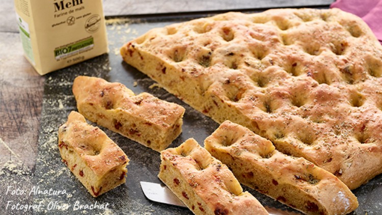 Ein rechteckiges, gebackenes Focaccia-Brot mit Kräutern liegt aufgeschnitten auf einer dunklen Oberfläche neben einem Mehlpaket.