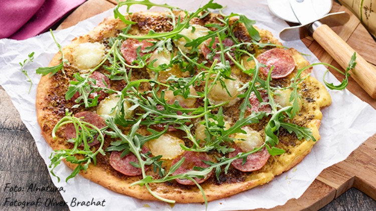 Knusprige Pizza mit grünem Rucola, Salami, geschmolzenem Käse und Kräutern auf Backpapier, daneben Pizzaroller und Holzbrett.
