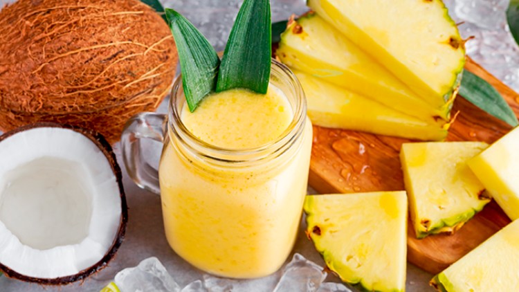 Gelber Smoothie in einem Glas mit Ananasblättern, umgeben von Ananasscheiben, Kokosnüssen, Limettenstücken und Eiswürfeln.