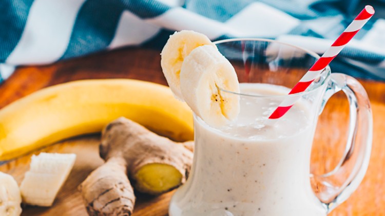 Glaskrug mit hellen Bananen-Smoothie, geschmückt mit Bananenscheiben und rot-weißem Strohhalm, daneben Banane und Ingwerknolle.