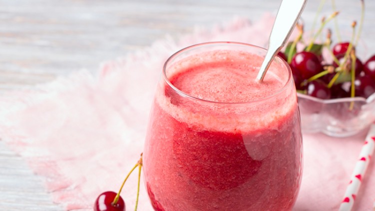 Ein Glas mit rotem Kirsch-Smoothie, Löffel darin, daneben frische Kirschen auf rosafarbenem Tuch und Herzensstrohhalme.