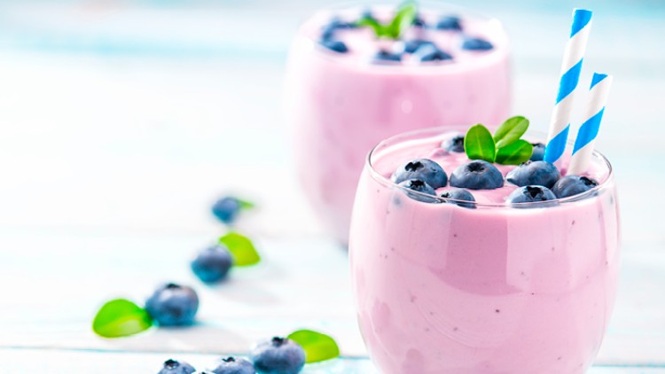 Zwei Gläser Blaubeersmoothie mit Blaubeeren, grünem Blattdekor und blau-weiß gestreiften Strohhalmen auf hellem Holz.