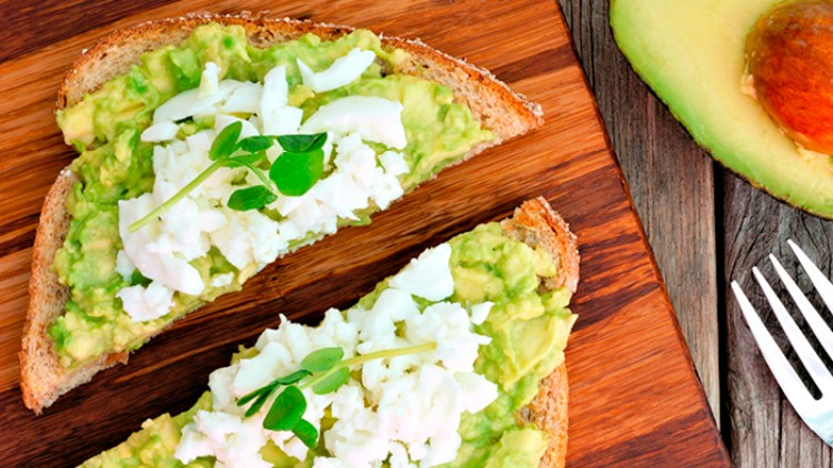 Zwei Scheiben Vollkornbrot mit Avocadocreme, zerbröseltem Ei und Kräutern auf einem Holzbrett, daneben eine halbe Avocado und eine Gabel.