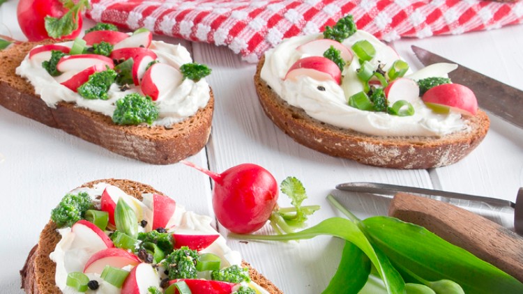 Drei Scheiben Vollkornbrot mit Frischkäse, Radieschen, Frühlingszwiebeln und Kräuterpesto auf weißem Holztisch angerichtet.