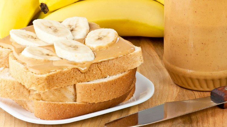 Ein Teller mit Sandwiches aus Weißbrot, Erdnussbutter und Bananenscheiben steht neben einem offenen Glas Erdnussbutter und Bananen.
