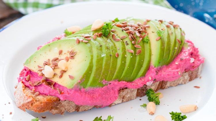 Scheibe Brot mit rotem Brotaufstrich, dünn geschnittener Avocado, Petersilie, Nüssen und Samen auf einem weißen Teller.