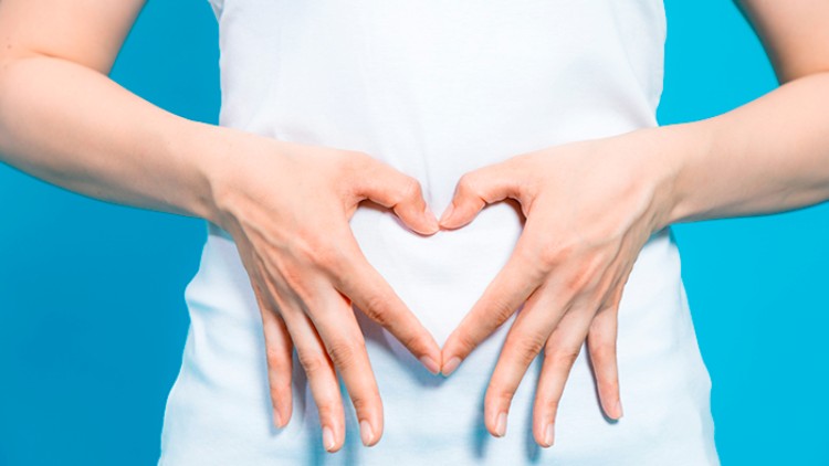 Zwei Hände formen ein Herz über dem Bauch, getragen wird ein weißes Shirt vor einem hellblauen Hintergrund.