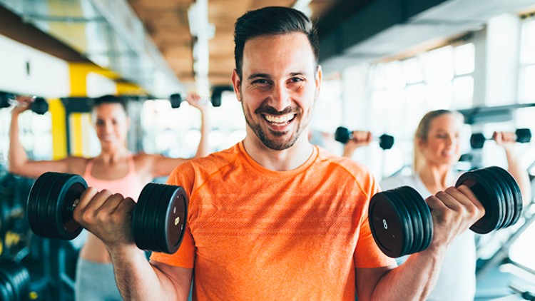 Person in orangefarbenem Shirt hebt zwei Hanteln im Fitnessstudio, im Hintergrund trainieren weitere Personen mit Hanteln.