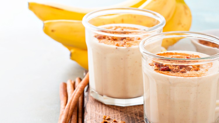Zwei Gläser mit cremigem Bananensmoothie, Zimt und Müsli, daneben Zimtstangen und frische Bananen auf einem Holzbrett.