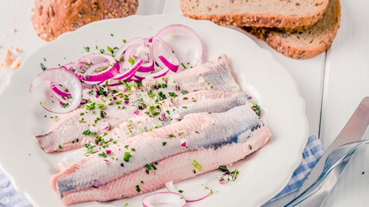 Drei marinierte Fischfilets mit roten Zwiebelringen und Kräutern auf einem weißen Teller, daneben Brot und Besteck.