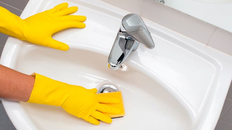 Zwei Hände in gelben Gummihandschuhen reinigen mit einem gelben Schwamm ein weißes Waschbecken mit Chromarmatur.