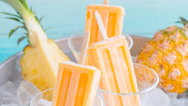 Drei gelbe Eis am Stiel in Gläsern mit Eiswürfeln, daneben eine frische Ananas und eine Ananasscheibe.