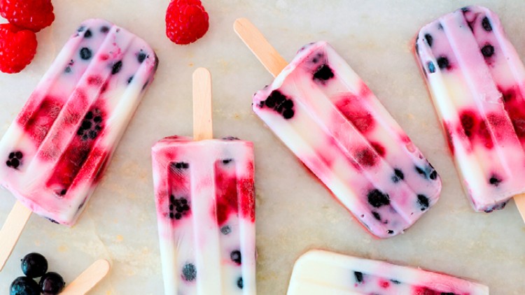 Fruchtige Eis am Stiel mit Joghurt, Himbeeren und Blaubeeren liegen auf einer hellen Oberfläche mit Holzstielen.