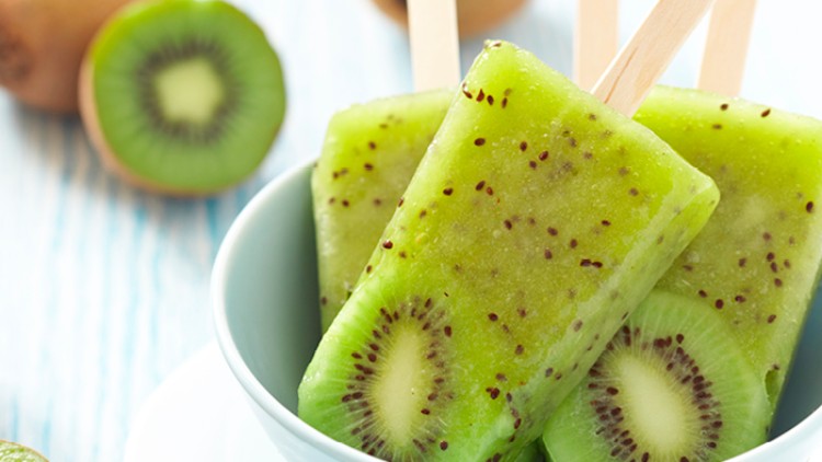 Drei grüne Kiwi-Eis am Stiel in einer Schale, daneben frische Kiwis und Kiwihälften auf hellem Untergrund.