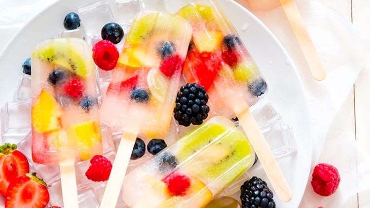 Durchsichtige Fruchteislollies mit Beeren, Kiwi- und Pfirsichstücken liegen auf Eiswürfeln auf einem weißen Teller.