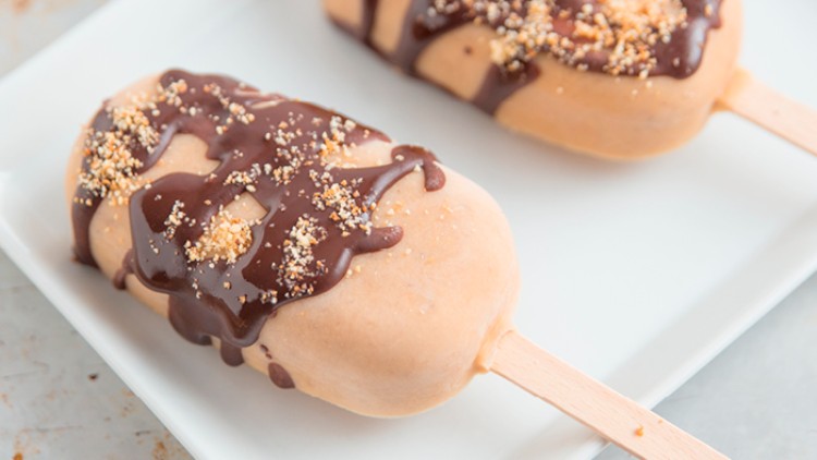 Zwei helle Eis am Stiel mit Schokoladensauce und Krokantstreuseln auf rechteckigem weißen Teller.
