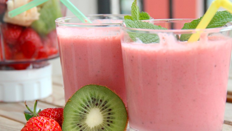 Zwei Gläser rosa Smoothie mit gelbem und grünem Strohhalm, daneben Erdbeeren, eine halbierte Kiwi und Dekoration mit Minze.