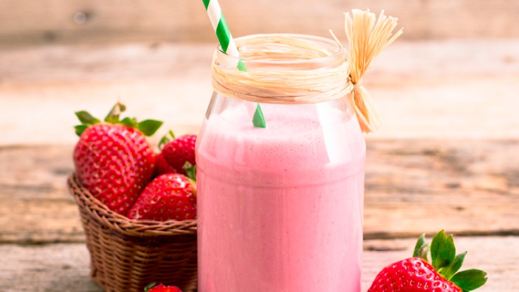 Ein Glas Erdbeermilchshake mit grün-weißem Strohhalm und Bastband steht neben frischen Erdbeeren und einem Korb auf Holz.