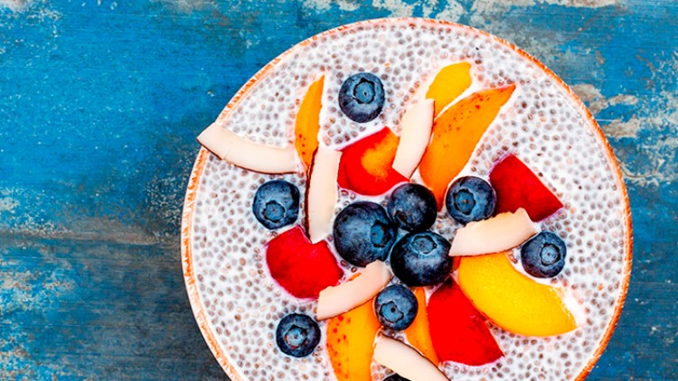 Eine Schale mit Chiasamenpudding, belegt mit Blaubeeren, Pfirsichstücken, Nektarine und Kokosflocken auf blauem Untergrund.