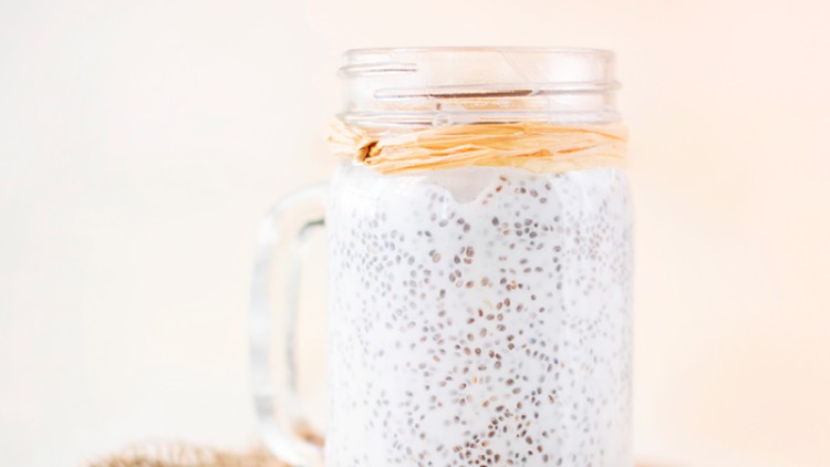 Ein Glas mit Henkel, gefüllt mit weißem Chia-Pudding und dekoriert mit Naturband, steht auf braunem Jutetuch.