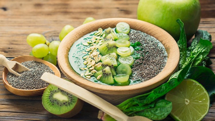 Holzschale mit grüner Smoothie-Bowl, dekoriert mit Kiwi, Chia-Samen, Haferflocken und Trauben auf Holzuntergrund.